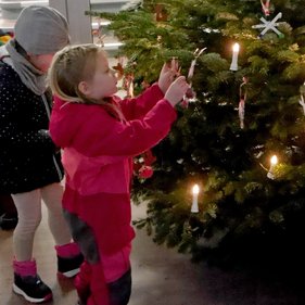 Baumschmuck 2023 Zwei Kinder in Winterkleidung schmücken einen Weihnachtsbaum, wobei das eine Kind, welches eine rosafarbene Jacke trägt, eine Dekoration berührt.