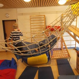 Kindertagesstätte Hinteres Sandfeld Zwei Kinder spielen in einem Raum mit Klettergerüsten und Hängematten aus Seilen. Ein Kind sitzt in der Hängematte und lächelt, während das andere Kind daneben steht. Der Raum hat eine Holzdecke und an den Wänden sind Kletterleitern befestigt. Auf dem Boden liegen weiche Matten in verschiedenen Farben.