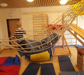 Zwei Kinder spielen in einem Raum mit Klettergerüsten und Hängematten aus Seilen. Ein Kind sitzt in der Hängematte und lächelt, während das andere Kind daneben steht. Der Raum hat eine Holzdecke und an den Wänden sind Kletterleitern befestigt. Auf dem Boden liegen weiche Matten in verschiedenen Farben.