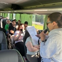Frau spricht mit einem Mikrofon zu einer Gruppe von Menschen in einem Bus.