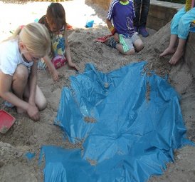 Mehrere Kinder spielen im Sandkasten und graben mit den Händen im Sand. Eine blaue Plane ist teilweise im Sand vergraben. Die Kinder tragen bunte Kleidung und sind konzentriert bei ihrer Tätigkeit.