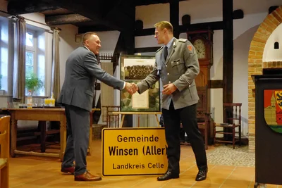 Bürgermeister Dirk Oelmann und Oberstleutnant Daniel Beer geben sich die Hand. Hinter ihnen steht eine gerahmte Urkunde, davor das Ortsschild „Gemeinde Winsen (Aller), Landkreis Celle“.