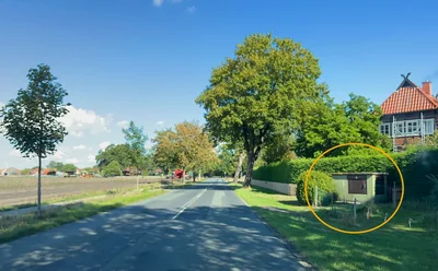 Straße in Wolthausen mit Bäumen und Wiese. Ein gelber Kreis markiert rechts am Straßenrand die Trafostation 13-446 Kraehenberg, die erneuert wird.