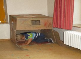 Ein Kind kriecht in eine große, bemalte Pappkiste, die als Spielhaus dient. Der Raum hat helle Wände und einen Heizkörper unter einem Fenster mit roten Vorhängen. Auf dem Boden liegen bunte Papierstücke verstreut.