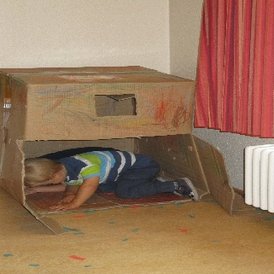 Kindertagesstätte Hinteres Sandfeld Ein Kind kriecht in eine große, bemalte Pappkiste, die als Spielhaus dient. Der Raum hat helle Wände und einen Heizkörper unter einem Fenster mit roten Vorhängen. Auf dem Boden liegen bunte Papierstücke verstreut.