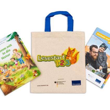 Ein Set zum Lesestart 1-2-3 Projekt, bestehend aus einem Buch mit dem Titel 'Komm mit in die Natur', auf dessen Cover eine Familie in einer Waldszene abgebildet ist, einer naturfarbenen Stofftasche mit blauen Tragegriffen und dem 'Lesestart 1-2-3' Logo, sowie einem Buch mit dem Titel 'Vorlesen und Erzählen' mit einem Foto von einem Vater, der sein Kind auf den Schultern trägt. Die Webseite www.lesestart.de ist unten auf der Tasche vermerkt, neben den Logos des Bundesministeriums für Bildung und Forschung und der Stiftung Lesen.
