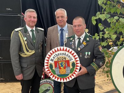 Drei Männer in Uniform posieren mit einer Plakette "Rathauskönig 2024".