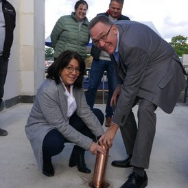 Grundsteinlegung Kindertagesstätte Südwinsen Zwei Personen, eine Frau im grauen Mantel und ein Mann im grauen Anzug, knien auf einem Betonboden und halten gemeinsam eine Kupferzylinder fest, der in den Boden eingesetzt wird. Im Hintergrund stehen drei weitere Personen und schauen zu
