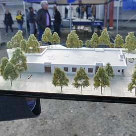 Grundsteinlegung Kindertagesstätte Südwinsen Das Bild zeigt ein Architekturmodell eines flachen, modernen Gebäudes auf einem Holzsockel. Das Gebäude ist von vielen kleinen Bäumen umgeben, die das Landschaftsdesign darstellen. Es gibt auch markierte Bereiche und Strukturen, die möglicherweise Wege und andere Gebäudeelemente darstellen. Das Modell wird von einer Hand gehalten und im Hintergrund sind unscharfe Figuren zu sehen, die sich auf einer Veranstaltung bewegen