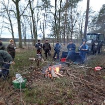 Gruppe von Menschen um ein Lagerfeuer im Wald, mit Ästen und einem Traktor im Hintergrund.