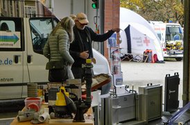 Zwei Personen betrachten technische Geräte in einer Garage, im Hintergrund ein Zelt mit rotem Kreuz.