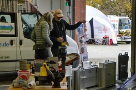 Hochwasseraktionstag Zwei Personen betrachten technische Geräte in einer Garage, im Hintergrund ein Zelt mit rotem Kreuz.