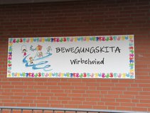 Schild mit der Aufschrift "Bewegungskita Wirbelwind" an einer roten Backsteinmauer.