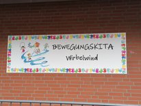 Neuer Name für die Kita Schild mit der Aufschrift "Bewegungskita Wirbelwind" an einer roten Backsteinmauer.
