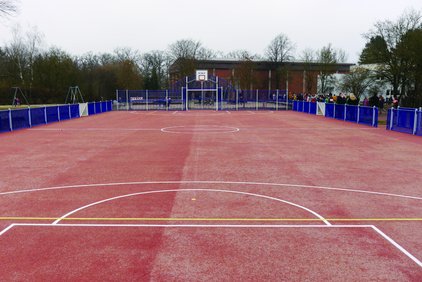 Leerer roter Sportplatz mit blauen Zäunen und einem Basketballkorb im Hintergrund.