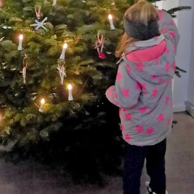 Baumschmuck 2023 Kind mit dem Rücken zum Betrachter steht, das einen Weihnachtsbaum mit echten Kerzen betrachtet. Das Kind trägt eine graue Winterjacke mit pinken Sternen und eine schwarze Mütze.