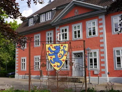 Das rote Rathaus der Gemeinde Winsen (Aller) mit dem farbigen Gemeindewappen im Vordergrund und einem Blumenbeet.
