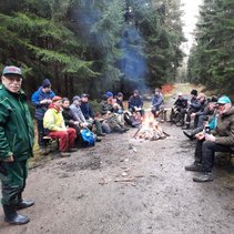 Gruppe von Menschen sitzt um ein Lagerfeuer im Wald, einige trinken, einer steht lächelnd.