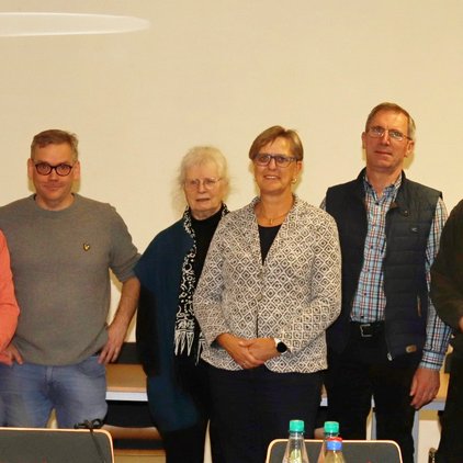 Eine Gruppe von acht Personen steht lächelnd vor einer weißen Wand.