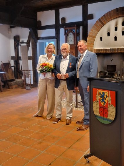 Drei Personen in historischer Kulisse; eine Person hält einen Blumenstrauß.