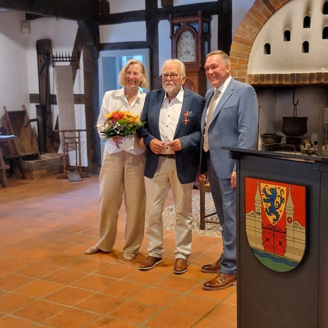Drei Personen in historischer Kulisse; eine Person hält einen Blumenstrauß.