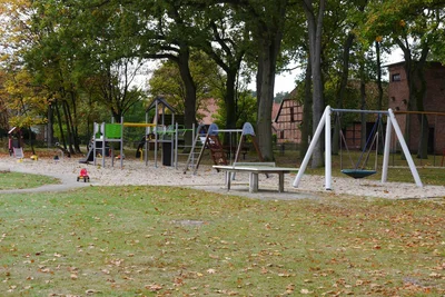 Blick über den Spielplatz in Stedden: Sandfläche mit Klettergerüst und Nestschaukel unter Bäumen, davor eine Rasenfläche.