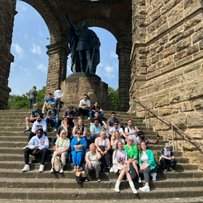 Bericht zur Bildungsreise mit Migranten zum Denkmal Kaiser Wilhelm Gruppe von Menschen sitzt auf Treppenstufen vor einer großen Statue.