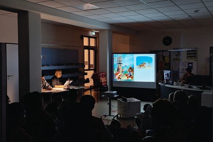 Dunkler Raum mit zwei Personen, die vorlesen, und einer Leinwand mit bunten Illustrationen