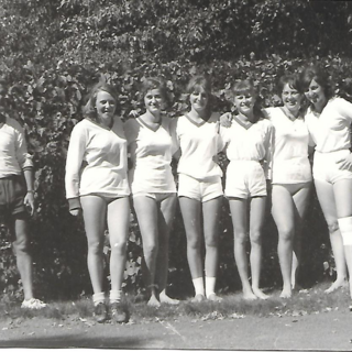 Schwarz-weiß-Foto von sechs Faustballspielerinnen in Sportkleidung mit ihrem Trainer, aufgenommen im Freien in den 1960er-Jahren.