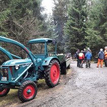 Blau-Grüner Traktor mit Anhänger auf Waldweg, dahinter Gruppe von Menschen im Gespräch.