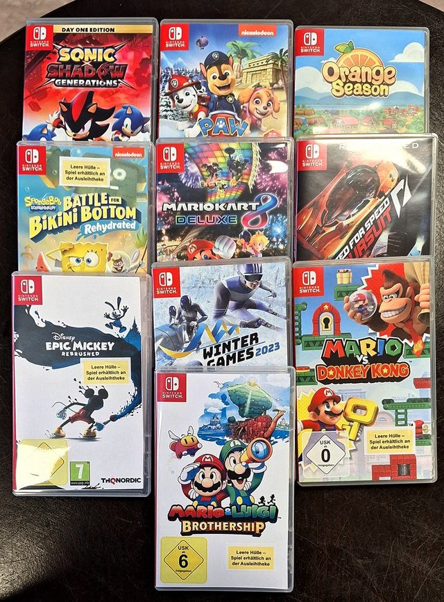 Nintendo Switch-Spielhüllen, darunter „Sonic Shadow Generations“, „Paw Patrol“, „Orange Season“, „SpongeBob Battle for Bikini Bottom Rehydrated“, „Mario Kart 8 Deluxe“, „Need for Speed Hot Pursuit“, „Disney Epic Mickey Rebrushed“, „Winter Games 2023“, „Mario vs. Donkey Kong“ und „Mario & Luigi: Brothership“. Manche Hüllen tragen gelbe Hinweise: „Leere Hülle – Spiel erhältlich an der Ausleihtheke