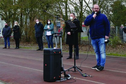 Ein Mann spricht in ein Mikrofon auf einem Sportplatz, im Hintergrund stehen Zuhörer.
