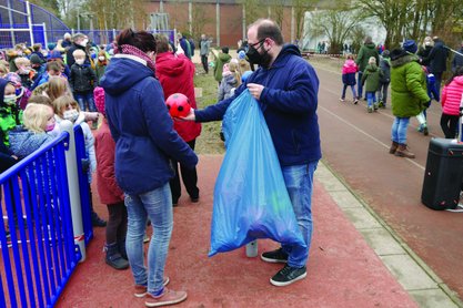 Mann verteilt aus einem blauen Sack rote Bälle an Kinder auf einem Sportplatz.