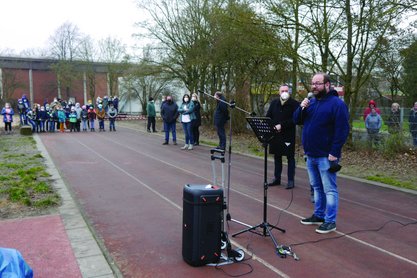 Ein Mann spricht in ein Mikrofon auf einem Sportplatz, Menschen im Hintergrund hören zu.