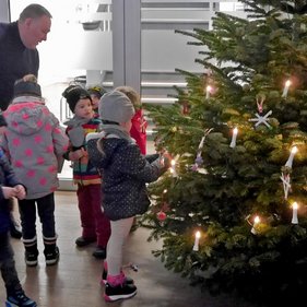 Baumschmuck 2023 Erwachsener und Kinder in Winterkleidung stehen um einen Weihnachtsbaum herum, der mit Kerzen und selbstgebastelten Dekorationen geschmückt ist.