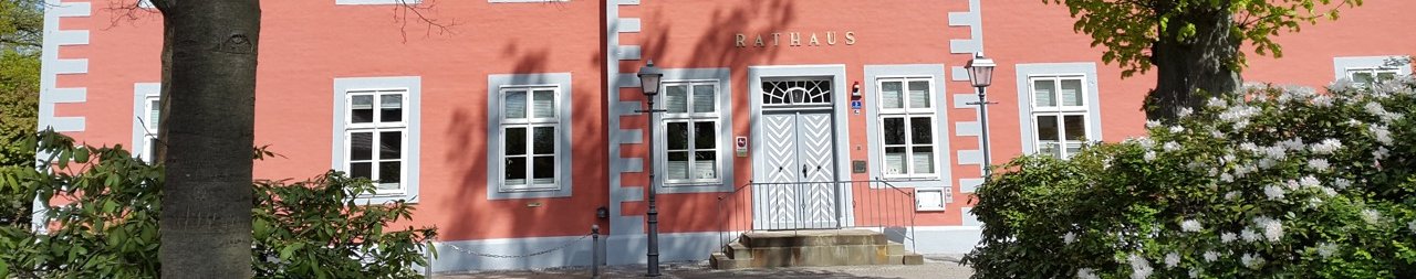 Ein farbenfrohes Rathausgebäude mit roter Fassade und weißen Fenstern. Über dem zentralen Eingang befindet sich ein dreieckiger Giebel mit einem Wappen. Vor dem Gebäude stehen zwei Laternen, und es ist von Bäumen und Büschen umgeben. Der Himmel ist klar mit wenigen Wolken.