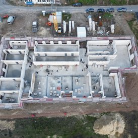 Grundsteinlegung Kindertagesstätte Südwinsen Ein Luftbild zeigt eine Baustelle, auf der mehrere Betonstrukturen im Bau sind. Die Strukturen sind von Gerüsten umgeben. Es sind Arbeiter auf der Baustelle zu sehen, die sich an verschiedenen Stellen bewegen. Mehrere Fahrzeuge, einschließlich eines Krans, sind auf der Baustelle und in einem angrenzenden Parkbereich geparkt. Neben der Baustelle befindet sich ein fertiggestelltes Gebäude mit einer grünen Rasenfläche.
