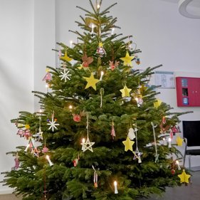 Baumschmuck 2023 Großer Weihnachtsbaum, geschmückt mit bunten Sternen, Kerzen und anderen handgefertigten Dekorationen in einem Innenraum.