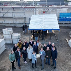 Grundsteinlegung Kindertagesstätte Südwinsen Eine Gruppe von Personen steht auf einer Baustelle, einige unter einem blauen Zelt, während andere sich neben gestapelten Baumaterialien versammeln. Im Hintergrund sind Gerüste und eine Wand mit dem Logo 'GOLDBECK' sichtbar.