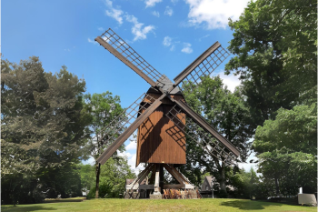 Alte Bockwindmühle mit dunkler Holzkonstruktion und vier großen Flügeln, umgeben von grünen Bäumen unter einem blauen Himmel mit vereinzelten Wolken.