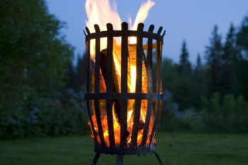  Eine Feuerschale mit lodernden Flammen in der Dämmerung, platziert auf einer grünen Wiese mit Bäumen im Hintergrund.