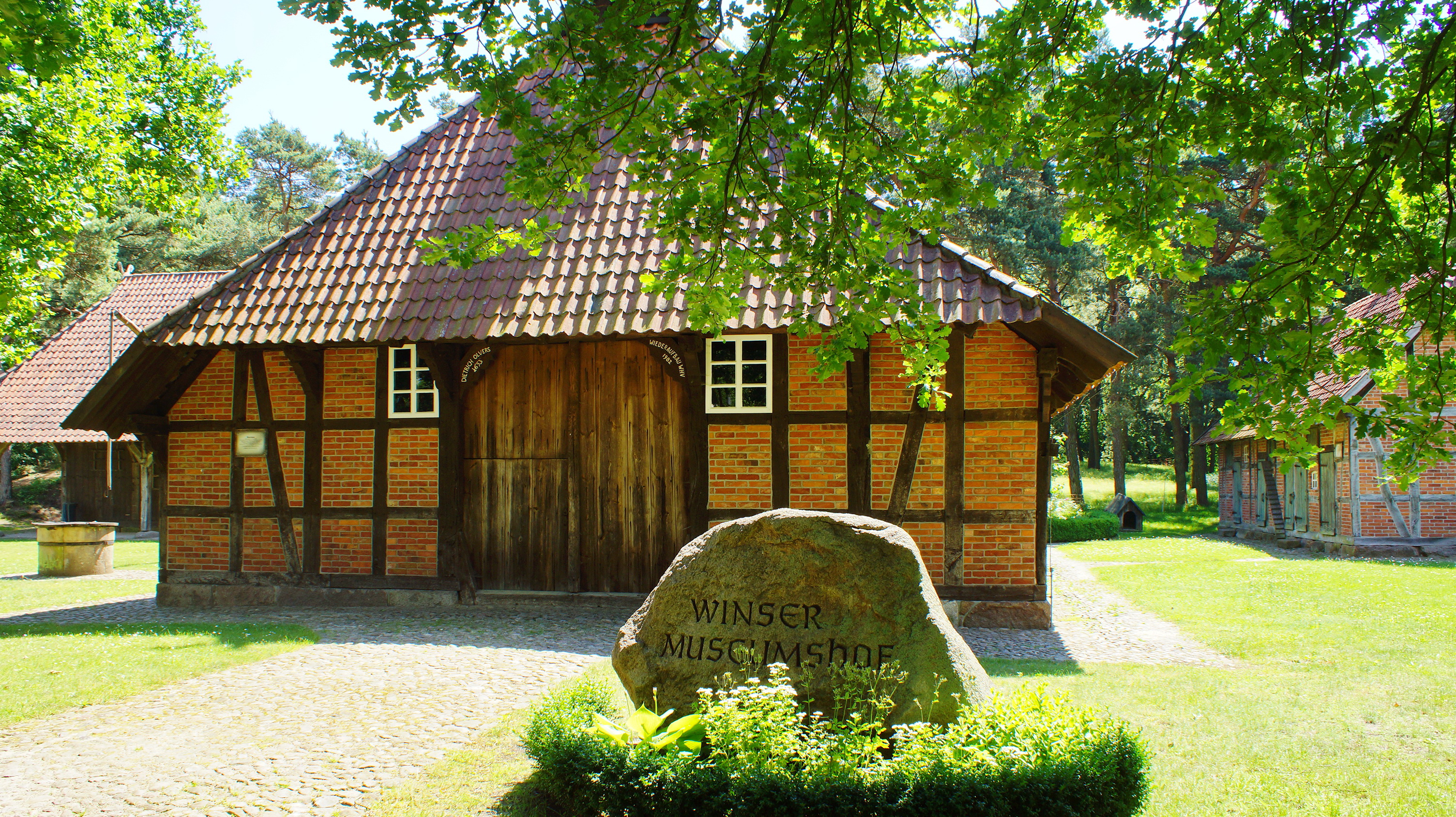 Fotografie eines traditionellen, niedersächsischen Bauernhauses im Freilichtmuseum, mit sichtbaren Fachwerkbalken und Ziegeln, vor dem ein großer Stein mit der Inschrift "Winsener Museumsdorf" steht, umgeben von grünen Bäumen und einem gepflegten Rasen.