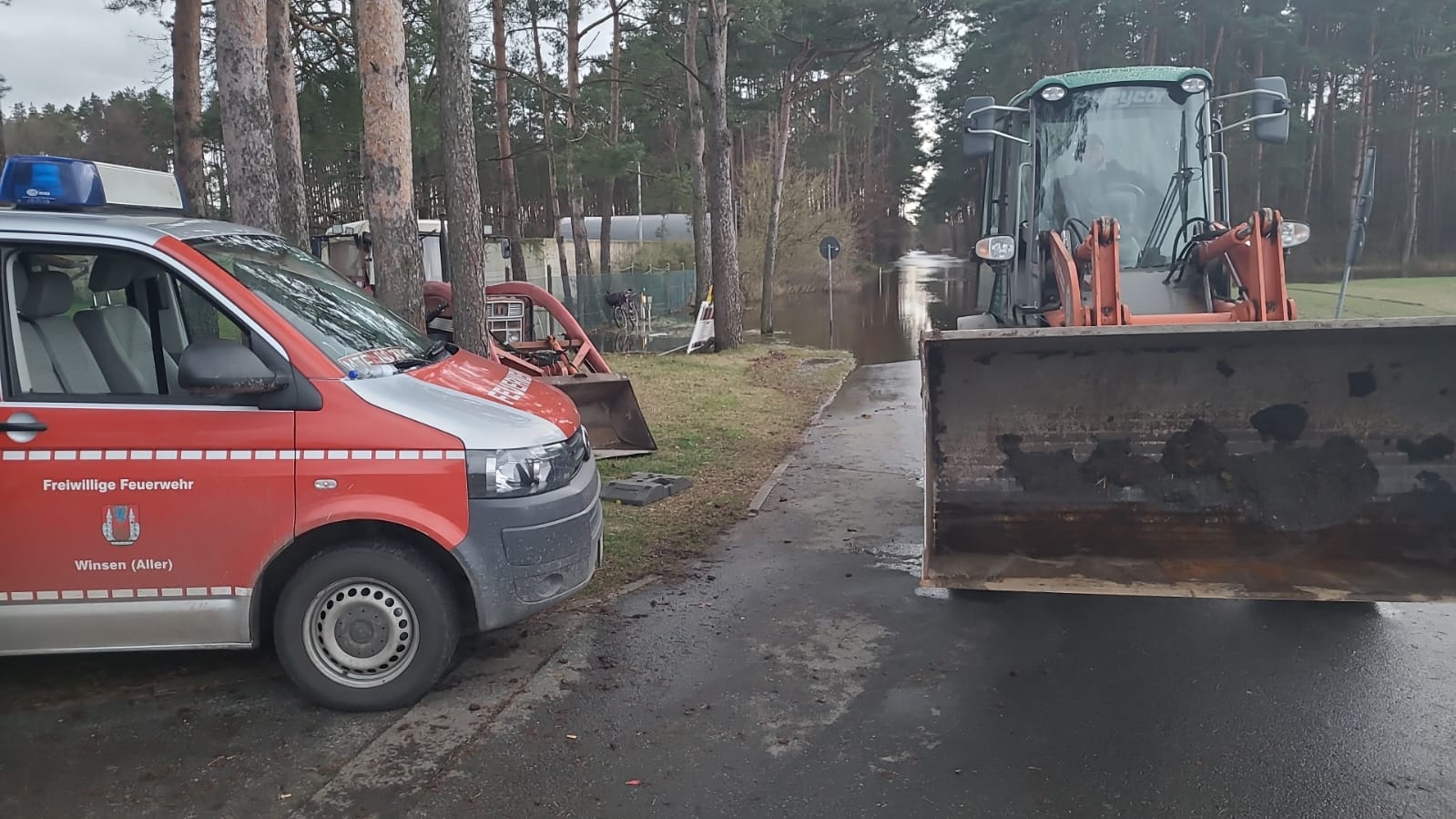 Feuerwehrfahrzeug und Radlader neben einer überfluteten Straße.