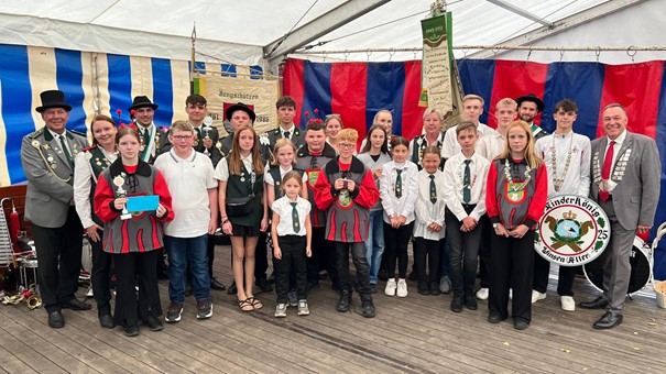 Gruppe in Festkleidung mit Auszeichnungen vor buntem Zelthintergrund.