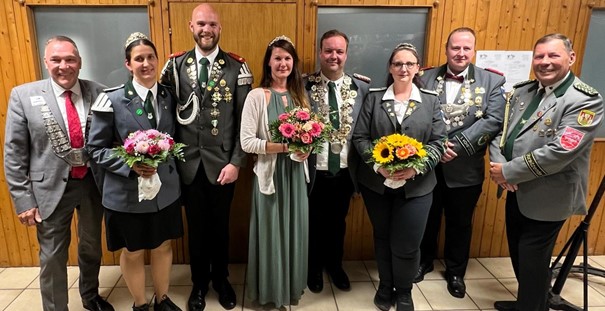 Acht Personen in Schützenuniform mit Blumensträußen vor Holzwand posierend.